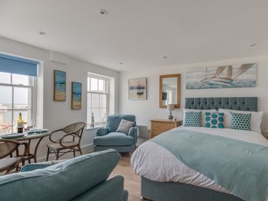 A bedroom with a bed armchairs a small dining table with chairs artworks on walls and windows at 6 The Beaches in Weymouth