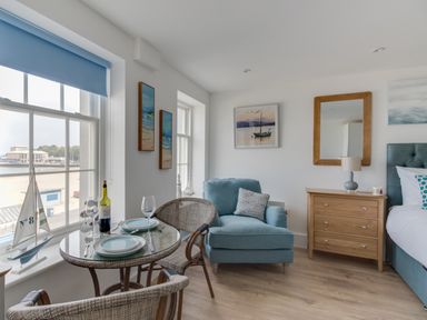A seating area with a blue armchair wooden chest of drawers glass table with two chairs and a window showing an outside view at 6 The Beaches in Weymouth