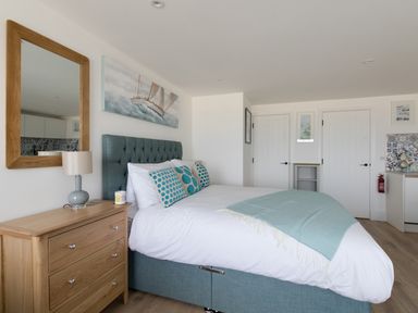 A bedroom with a teal upholstered bed a wooden dresser with a lamp and mirror and two white doors at 6 The Beaches in Weymouth