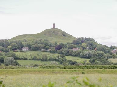 A grassy hill with a stone tower on top and trees with houses at the base