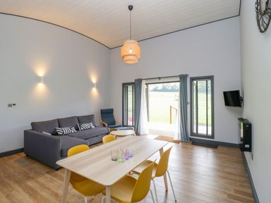 A living room with a dining table and yellow chairs a gray sofa a blue armchair a wall-mounted TV and large windows at Bluebell in Sticklinch West Pennard Glastonbury
