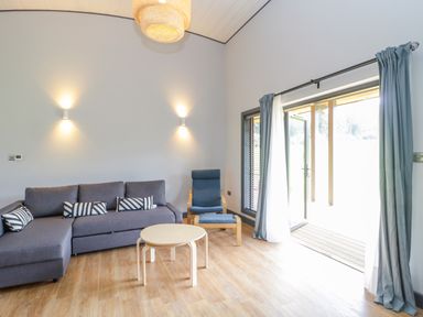 A living room with a gray sectional sofa and a wooden armchair next to a sliding glass door with blue curtains at Bluebell in Sticklinch West Pennard Glastonbury