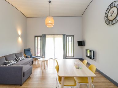 A living room with a gray sofa a blue armchair a wooden dining table with yellow chairs a round coffee table a wall clock and a wall-mounted TV at Bluebell in Sticklinch West Pennard Glastonbury