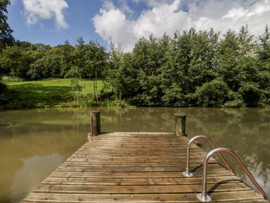 A wooden dock with metal handrails extending over a small pond surrounded by trees and grassy area at Bluebell in Sticklinch West Pennard Glastonbury