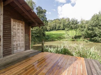 A wooden cabin with a deck beside a pond surrounded by grass and trees at Bluebell in Sticklinch West Pennard Glastonbury
