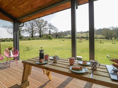 A covered outdoor patio with a wooden picnic table set for breakfast and a grassy field with trees in the background at Bluebell in Sticklinch West Pennard Glastonbury