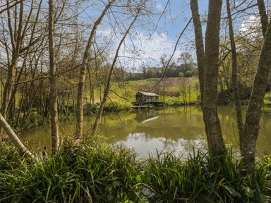 A small wooden cabin by a pond surrounded by trees and grassy hills at Bluebell in Sticklinch West Pennard Glastonbury
