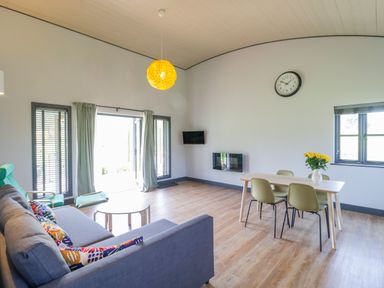 A living room with a grey sofa and colorful cushions a green chair and footstool a dining table with four chairs and yellow flowers at Primrose in Sticklinch West Pennard Glastonbury