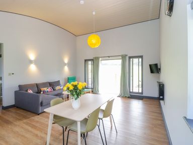 A dining table with flowers in a vase and chairs in a living room with a sofa and a wall mounted TV at Primrose in Sticklinch West Pennard Glastonbury