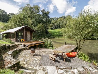 A small wooden cabin next to a pond with a wooden dock and a picnic table with benches at Primrose in Sticklinch West Pennard Glastonbury