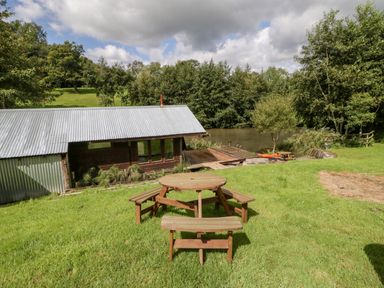 A grassy area with wooden picnic tables near a pond and a building with a corrugated metal roof at Primrose Sticklinch West Pennard Glastonbury