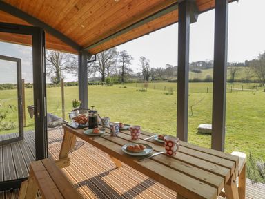 A covered wooden patio with a picnic table set with croissants and mugs overlooking a grassy field at Primrose in Sticklinch West Pennard Glastonbury