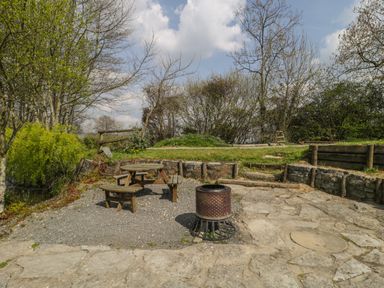 An outdoor seating area with a wooden picnic table and a metal fire pit surrounded by trees and grass at Primrose in Sticklinch West Pennard Glastonbury