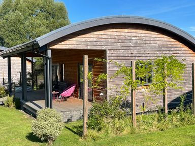 A wooden cabin with a curved roof and a porch with two pink chairs and a small table surrounded by greenery at Primrose in Sticklinch West Pennard Glastonbury