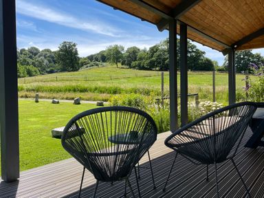 A covered wooden deck with two black wire chairs and a table overlooking a grassy field and trees at Primrose in Sticklinch West Pennard Glastonbury