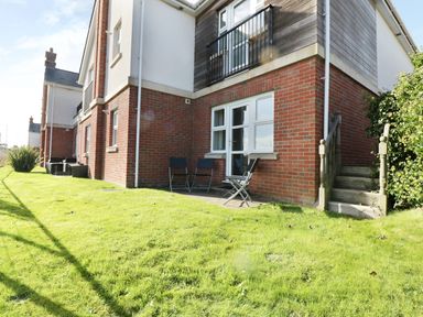 The exterior of a brick and wood house with a small patio and three chairs on grass at Bay View in Benllech