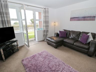 A living room with a leather sofa with purple and white cushions a purple rug a television on a stand a floor lamp and an open door leading outside at Bay View in Benllech