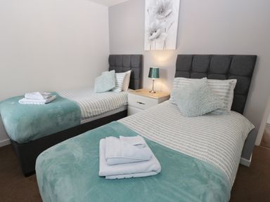 A bedroom with two single beds with teal throws and pillows and a nightstand with a lamp between at Bay View in Benllech