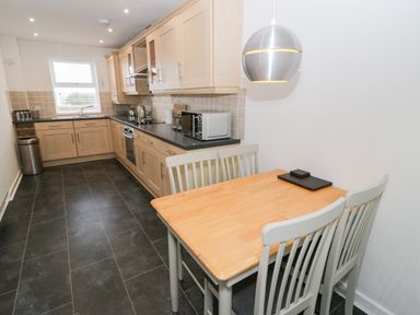 A kitchen with wooden cabinets a black countertop a dining table with four chairs and a silver hanging light at Bay View in Benllech