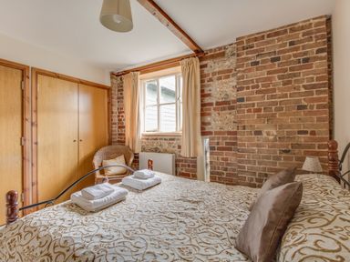 A bedroom with a bed covered in patterned bedding two folded towels on the bed a wicker chair by a window with curtains exposed brick walls and wooden closets at Manor Farm Barn in Dewlish