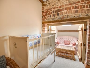 A small room with a white baby crib a pink couch with cushions and a wicker table at Manor Farm Barn in Dewlish