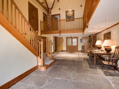 A hallway with a wooden staircase stone floor a rug wooden table with lamps and chairs at Manor Farm Barn in Dewlish