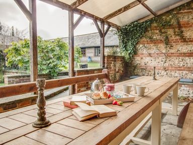 A covered outdoor dining area with a wooden table set with books mugs a tray of scones and strawberries at Manor Farm Barn in Dewlish
