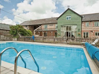 An outdoor swimming pool with metal handrails and wooden fence surrounding a brick and wood house at Manor Farm Barn in Dewlish