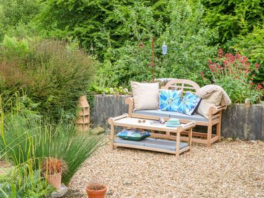 An outdoor seating area with a wooden bench and coffee table surrounded by plants at Woodstock Cottage in Birchill near Chardstock