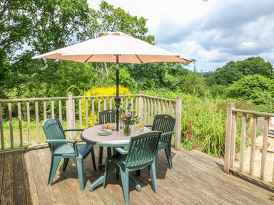 An outdoor wooden deck with a round table and four chairs under a large patio umbrella surrounded by greenery at Woodstock Cottage in Birchill near Chardstock