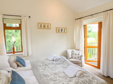 A bedroom with a bed pillows and folded towels near a window and an open door with a chair and cushions at Woodstock Cottage in Birchill near Chardstock