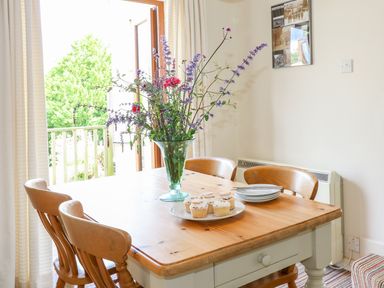 A dining room with a wooden table and chairs with flowers in a vase and cupcakes on a plate at Woodstock Cottage in Birchill near Chardstock