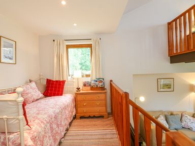 A small room with a daybed with cushions a wooden chest of drawers a window with curtains and a wooden railing overlooking a lower living area at Woodstock Cottage in Birchill near Chardstock