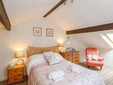 A bedroom with a bed two bedside tables and lamps a wooden dresser and a coral armchair at Woodstock Cottage in Birchill near Chardstock