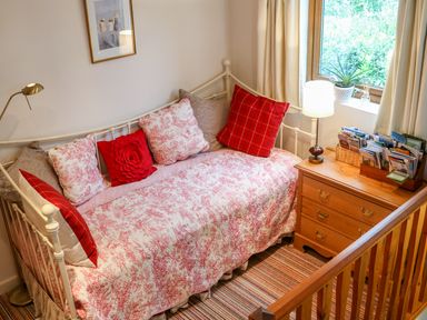 A bedroom with a bed with red and white cushions next to a wooden chest of drawers with a lamp and window at Woodstock Cottage in Birchill near Chardstock