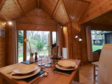 A wooden dining area with a table set for two near large windows overlooking a garden at Woodpeckers in Willand