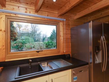 A kitchen with a black countertop sink a window showing trees and bushes and a stainless steel refrigerator at Woodpeckers in Willand