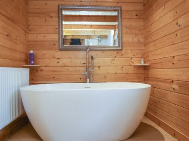 A bathroom with a white bathtub a mirror on a wooden wall and shelves with toiletries at Woodpeckers in Willand