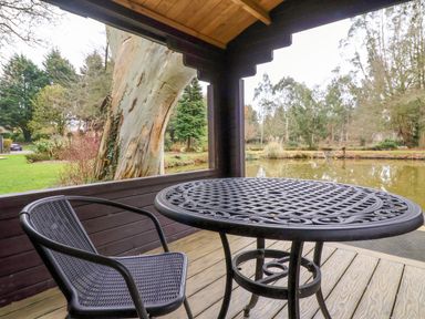 A covered wooden deck with a metal table and chair overlooking a pond and trees at Woodpeckers in Willand