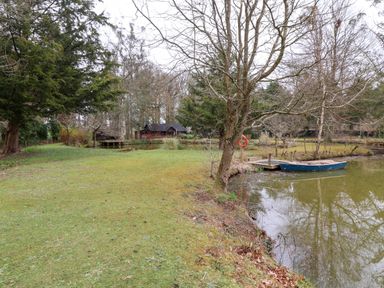 A pond with a small dock and a blue rowboat next to grass and trees at Woodpeckers in Willand