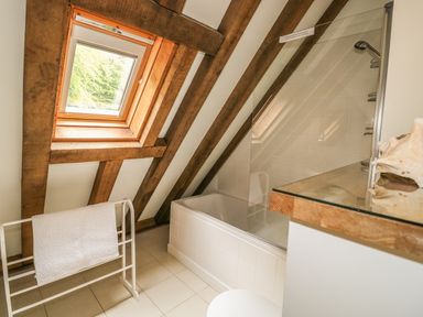 A bathroom with slanted wooden beams a bathtub with a glass shower screen a towel rack and a window at Trevinny Lodge No 37 Davidstow near Boscastle