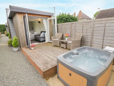 A small wooden cabin with an open door leading to a bedroom a wooden deck with two wicker chairs and a table a hot tub next to the deck and a gravel pathway at Alstroemeria Bretforton near Evesham