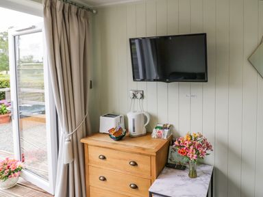 A wooden dresser with a toaster kettle and tea bags a TV on the wall and flowers on a small table near a balcony door at Alstroemeria Bretforton near Evesham