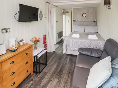 A studio room with a bed two pillows and folded towels a sofa with pillows a wooden chest of drawers and a wall mounted television at Alstroemeria Bretforton near Evesham