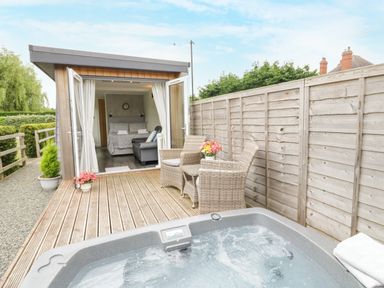 An outdoor deck with a hot tub and wicker chairs in front of a small building with open doors showing a bedroom at Alstroemeria in Bretforton near Evesham