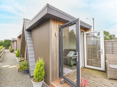 An outdoor wooden patio with open glass doors leading to a small building and potted plants along a gravel path at Alstroemeria Bretforton near Evesham