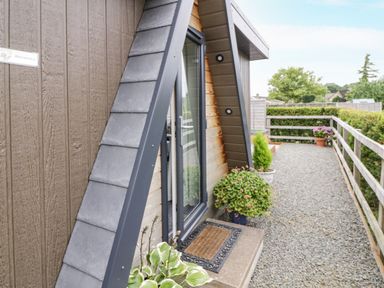 The exterior entrance of a wooden house with an A-frame design and gravel path with potted plants at Alstroemeria in Bretforton near Evesham