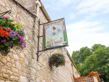 A stone building with a hanging pub sign and flower baskets outside at Pheasant Cottage in Priston Village BATH
