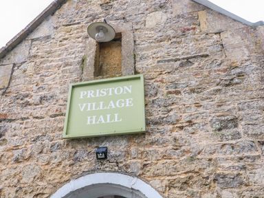 A stone building wall with a green sign reading Priston Village Hall and a light above the sign at Pheasant Cottage in Priston Village BATH