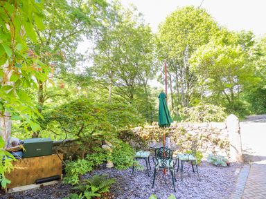 An outdoor seating area with metal chairs and table under a closed green umbrella surrounded by trees and plants at The Old Coach House in Llanarmon-Yn-Ial